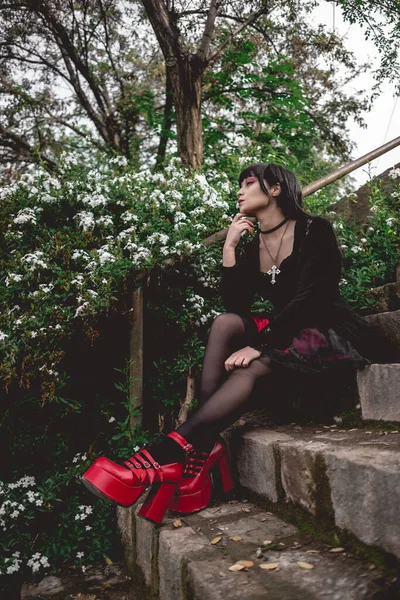 若いですとスキニーHispanicゴス女の子とともに黒ドレスと赤プラットフォームの靴で石の階段に座っている森の中の白い花の横に — ストック写真