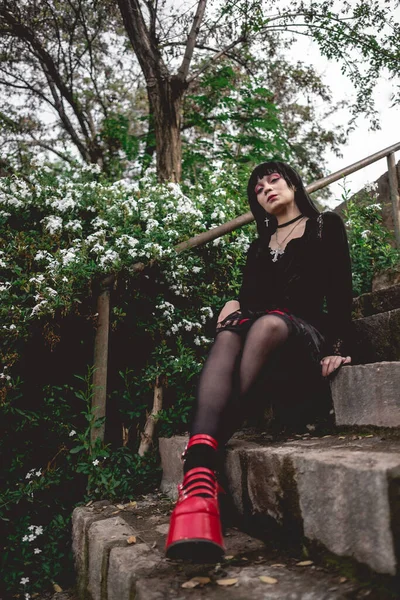 若いですとスキニーHispanicゴス女の子とともに黒ドレスと赤プラットフォームの靴で石の階段に座っている森の中の白い花の横に — ストック写真
