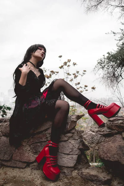 若いですとスキニーHispanicゴス女の子とともに黒ドレス パンストと赤プラットフォームの靴で小さな石の壁に座って森の中 — ストック写真