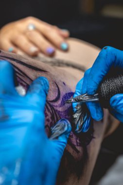 Dövmeci elleri mavi eldivenli, dövme makinesiyle, büyük bir kaplanın kalçasında ve güzel bir kadının kalçasında.