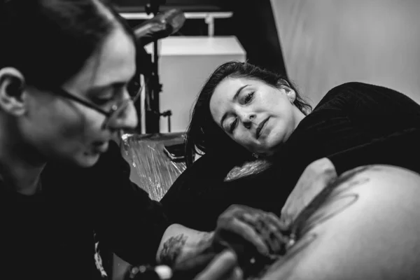 Concentrated young tattoo artist girl with glasses making a tattoo, with tattoo machine, of a big tiger, in the thigh and hips of a beautiful woman (in black and white)
