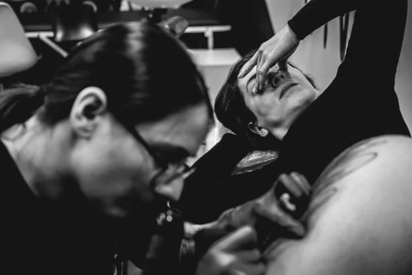 Concentrated young tattoo artist girl with glasses making a tattoo, with tattoo machine, of a big tiger, in the thigh and hips of a beautiful woman enduring pain (in black and white)