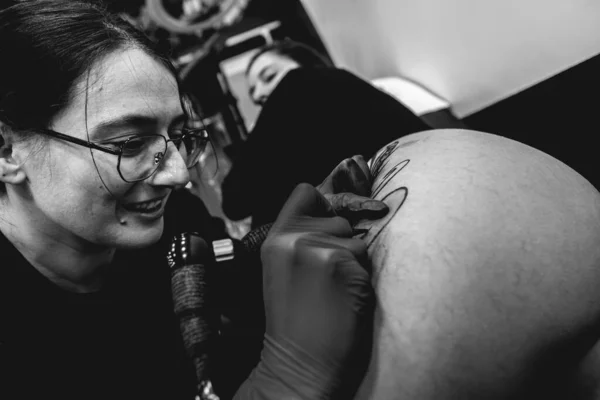 Smiling young tattoo artist girl with glasses making a tattoo, with tattoo machine, of a big tiger, in the thigh and hips of a beautiful woman (in black and white)
