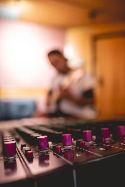 スタジオでいくつかのノブを持つサウンドミキサーボードの前でエレキギターを演奏する若い音楽家の男 — ストック写真