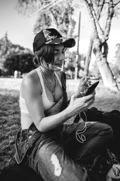 Rude, sexy and tattooed young girl with cap, denim dungarees and white crop top smiling and making a video call on the phone in the park (in black and white)