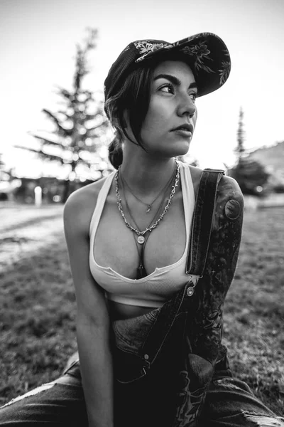 Rude, sexy and tattooed young girl with cap, denim dungarees and white crop top seated in the park in a sunny day (in black and white)