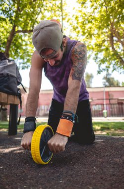 Healthy and fit young latin man with tattooed upper arm and shoulder, cap and purple shirt doing abdominal exercises with a ab roller wheel on a street workout park in a sunny day