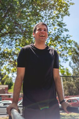 Very happy, healthy and fit young latin man with black shirt laughing and doing a calisthenic exercise routine (performs dips on parallel bars) in an street workout park in sunny day