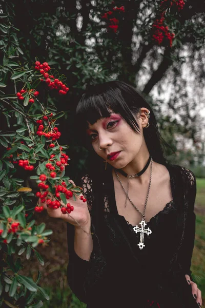 Young and skinny white goth girl with long black hair and black dress and pantyhose between big rowan berries trees in the forest
