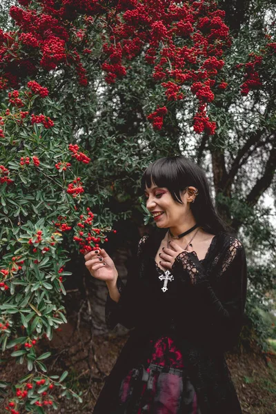 Young and skinny white goth girl with long black hair and black dress happy and laughing between big rowan berries trees in the forest