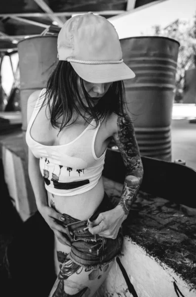 Rude, sexy and tattooed young girl with white crop top, denim shorts, cap and in his skateboard table in front of metal barrels (in black and white)