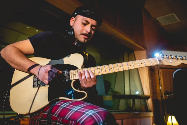 Young rocker musician with biker hat playing a cream electric guitar in a music studio