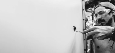 Fit young latino worker with PPE (work glasses) putting a screws into holes with screwdriver in a steel door of a electronic area (panoramic with big copy space in black and white)