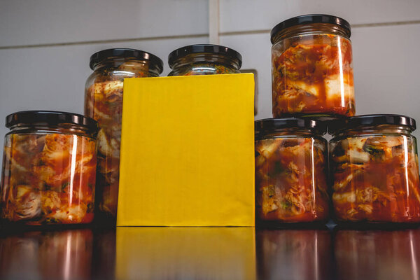 Пряный традиционный кимчи в стеклянных банках и желтая доска с копировальным местом на кухне (кимчи: смесь маринованных и ферментированных овощей, чеснок, перец чили, соль) 