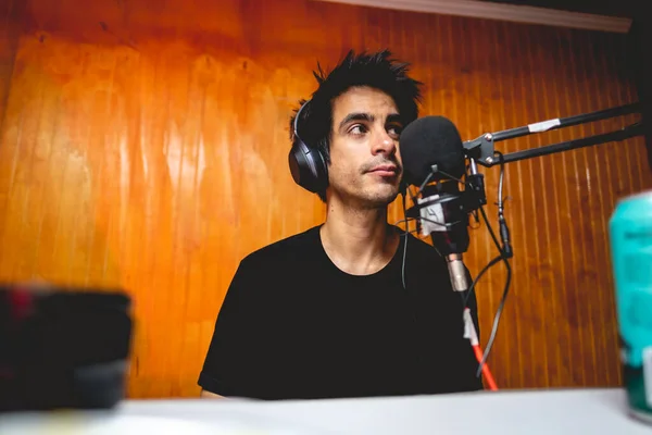 Young white man with black hair, headphones and black t-shirt in front of microphone recording in radio studio with wooden wall background