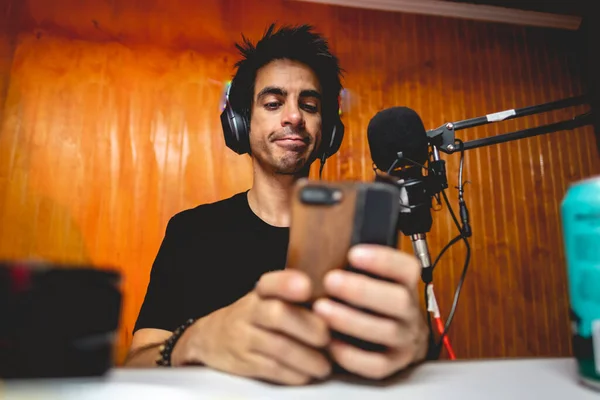 Young white man with black hair, headphones and black t-shirt in front of microphone using his phone in radio studio with wooden wall background