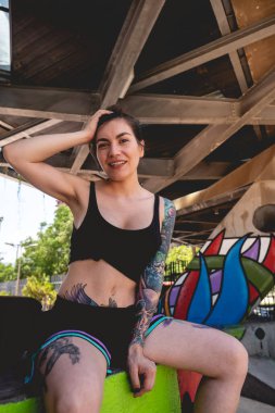 İsyancı stili: Siyah bluzlu ve kısa boylu seksi genç kadın, dövmeli ve kaykaylı şehir kaykayı parkında grafiti ile. 