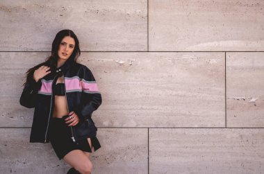 Şehir modası: genç esmer model deri ceket giyer ve sabah güneşli büyük taş bir binada uzun botlar giyer.