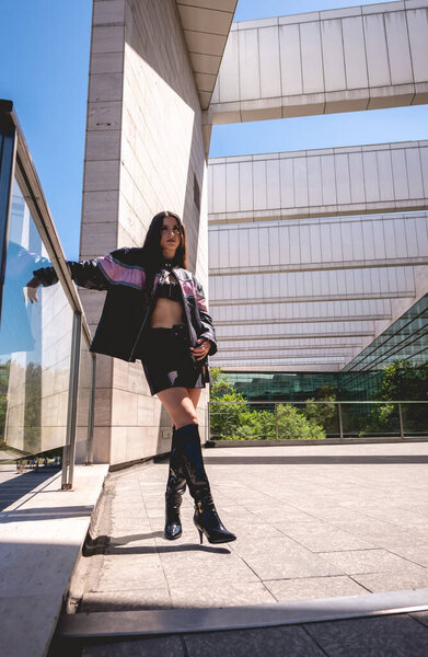 Urban chic: young brunette model wears leather jacket and long boots in public park with morning sunlight