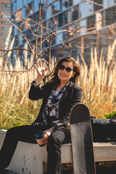 Modern, beautiful and young executive: sophisticated woman with sunglasses, coffee cup and skateboard, happy seated at urban park with fox tails and buildings in the back