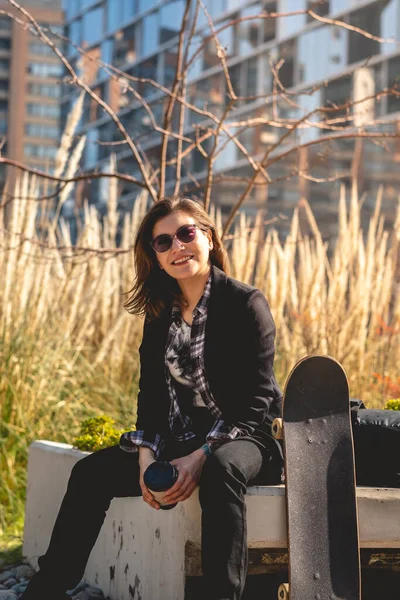 Modern, beautiful and young executive: sophisticated woman with sunglasses, coffee cup and skateboard, happy seated at urban park with fox tails and buildings in the back