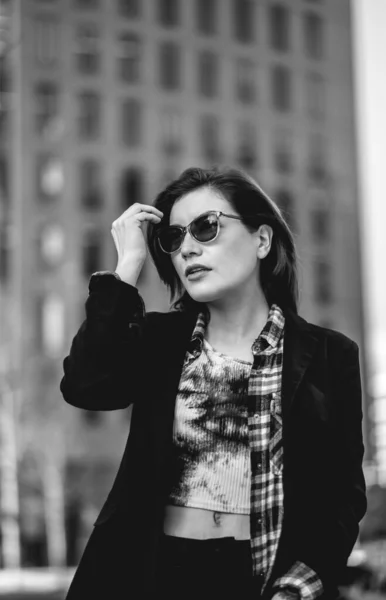 Modern, beautiful and young executive: sophisticated woman with sunglasses and blazer, with street and building behind (in black and white)