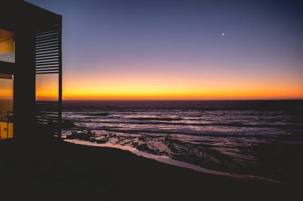Oceanic tranquility: silhouette of house in the cliff with warm lights and white crescent moon illuminating the dark purple ocean, as waves gently crash onto the beach sand, under a serene sunset sky
