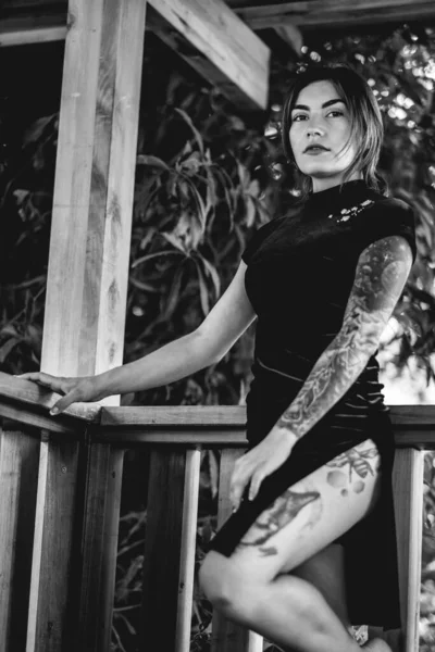 Exotic elegance: tattooed sexy young brunette woman in a blue chinese dress in a wooden pavilion surrounded by jungle (in black and white)