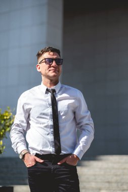 Profesyonel, çekici ve kendine güvenen beyaz gömlekli ve siyah kravatlı genç iş adamı şehir parkında güneşin altında duruyor.