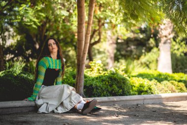 Doğa ve Latin güzelliği: Parkta yeşil doğa ile güneş ışınları arasında yeşil örülmüş süveter giyen genç esmer model