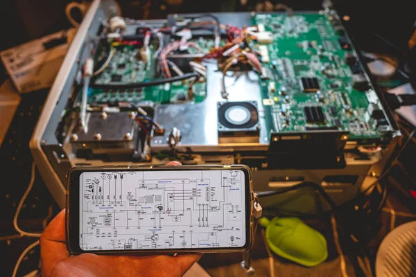 Elektronik diyagramı olan bir telefonu tutan: bilgisayar panosunun donanımı, devreleri ve bileşenlerini inceleyen