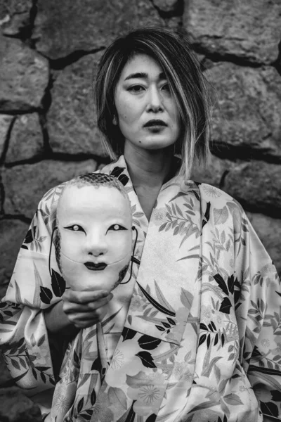 Japanese beauty: asian blonde-haired woman in traditional silk kimono with leaves design and Onnamen mask, in a japanese castle wall (in black and white)