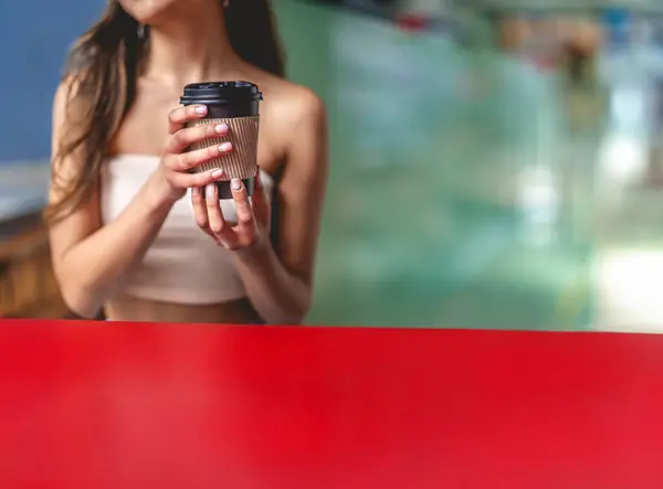 Urban lifestyle, coffee break and copy space: Young and beautiful latino girl with long and dark hair and beige top, holding an eco-friendly paper cup with coffee at a red table and big copy space