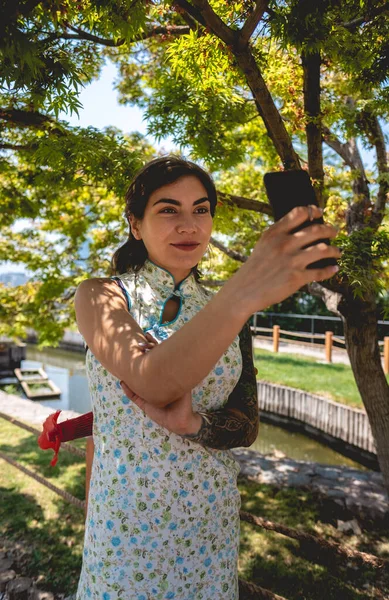 Technology and beauty japanese style: brunette girl with tattoos, asian dress, and phone making a selfie in a japanese garden