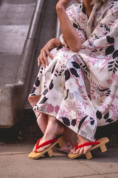 Japanese beauty, elegance and tradition: beautiful japanese model in traditional white silk kimono with pink leaves design and \'geta\' wooden sandals in a urban park