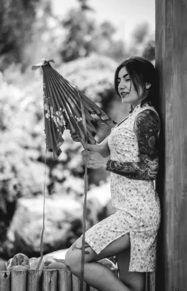Stunning beauty and japanese style: brunette girl with tattoos, asian dress, and paper umbrella, delighting in a sunny day with clear sky in a japanese garden (in black and white)