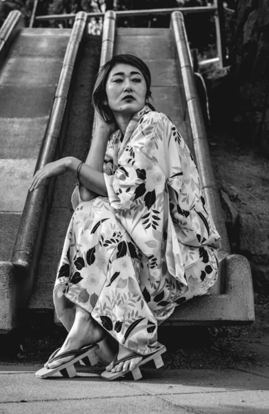 Japanese beauty, elegance and tradition: beautiful japanese model in traditional silk kimono with leaves design and \'geta\' wooden sandals in a urban park (in black and white)