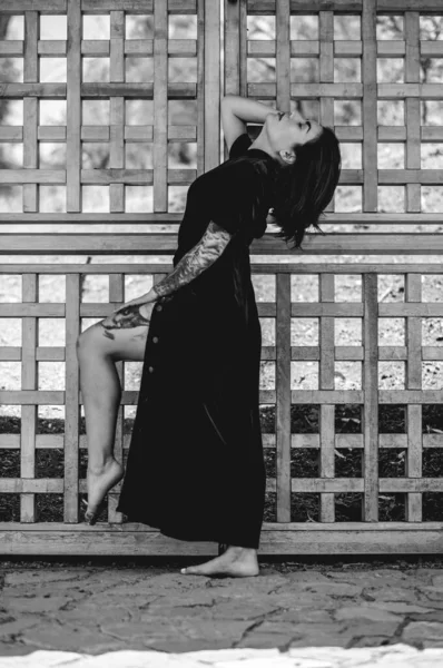 Exotic seduction: tattooed sexy young latina brunette woman in a crimson silk chinese dress in front of a wooden door in public park (in black and white)