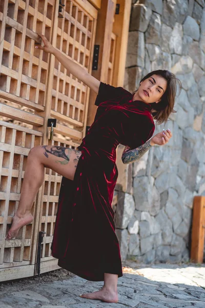Exotic seduction: tattooed sexy young latina brunette woman in a crimson silk chinese dress in front of a wooden door in public park