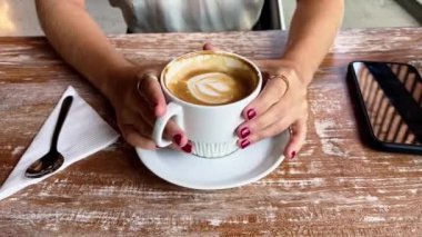 Sıradan kahve molası ayrıntıları: ahşap bir masada beyaz bir fincan kahve tutan kırmızı tırnaklı güzel kadın ellerine yakın çekim