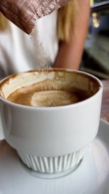 Sıradan kahve molası ayrıntıları: Bir kızın ahşap bir masada beyaz bir fincan kahveye şeker dökmesine yakın çekim