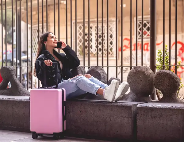 Stylish travel and urban exploration: vibrant young latina brunette with fashion look and pink suitcase making a call on the phone and exploring city downtown