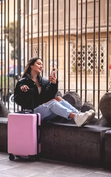 Stylish travel and urban exploration: vibrant young latina brunette with fashion look and pink suitcase making a video call on the phone and exploring city downtown