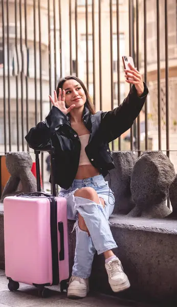 Stylish travel and urban exploration: vibrant young latina brunette with fashion look and pink suitcase making a video call on the phone and exploring city downtown