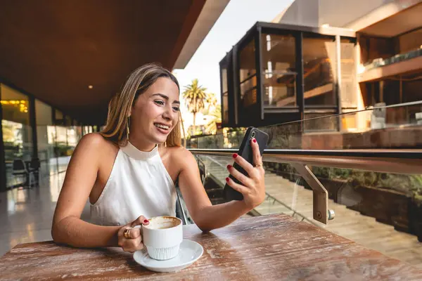 Casual business video meeting in a outdoor city coffee: beautiful blonde latina executive sipping coffee and having a video meeting in his phone
