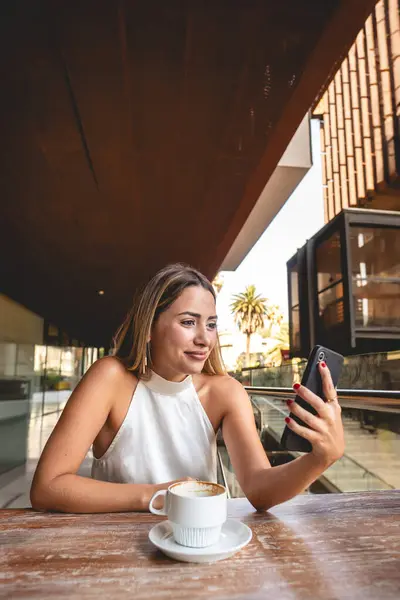 Casual business video meeting in a outdoor city coffee: beautiful blonde latina executive sipping coffee and having a video meeting in his phone