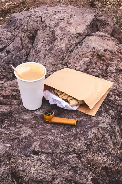 Relaxing roadside picnic snack: coffee, sandwich, and cannabis pipe set on a rock for a tranquil travel break