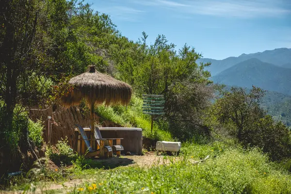 Hot tub and chairs for resting with straw canopy umbrella on a green hill with view of valley and mountains in a summer day