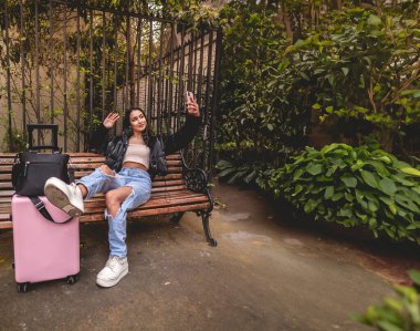 Şık ve genç gezgin Latin görünümlü esmer ve pembe valizli şehir parkını gezerken telefon kullanıyor.