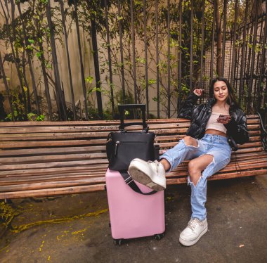 Şık ve genç gezgin Latin görünümlü esmer ve pembe valizli şehir parkını gezerken telefon kullanıyor.